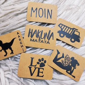 Può includere: Sei carte di carta marrone con testo e immagini nere. Le carte presentano le parole "Moin", "Hakuna Matata", "AARGH!" e "LOVE" con un'impronta di zampa. Le carte presentano anche una silhouette di dinosauro, un camion ribaltabile e una fata.