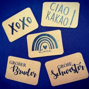 Può includere: Quattro carte marroni con testo nero. Le carte dicono "XOXO", "Ciao Kakao!" , "Grober Bruder", e "Grobe Schwester". Una carta ha un arcobaleno nero con un piccolo cuore e la parola "Love" sotto.