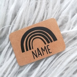 Può includere: Un adesivo rettangolare color kraft con un design ad arcobaleno nero e la parola "NAME" stampata sotto. L'adesivo è posto su uno sfondo di soffice pelliccia sintetica bianca.