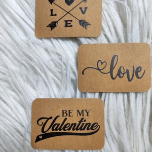 Può includere: Tre etichette regalo in carta marrone con testo nero a tema San Valentino. La prima etichetta ha un cuore e frecce con le lettere L, V ed E. La seconda etichetta dice "love" con un cuore. La terza etichetta dice "Be My Valentine" con un cuore.