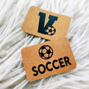 Könnte beinhalten: Zwei braune Papierkarten mit schwarzem Text und einer Fußballgrafik. Die erste Karte sagt "V" mit einem Fußball. Die zweite Karte sagt "SOCCER" mit einem Fußball.