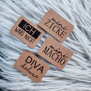 Può includere: Quattro etichette rettangolari marroni con testo nero. Le etichette recitano: "ICH WARS NICHT", "mini ZICKE", "mini MACHO" e "DIVA deluxe". Le etichette sono disposte su una superficie bianca e soffice.