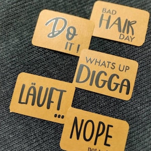 Può includere: Cinque adesivi rettangolari marroni con testo nero. Gli adesivi dicono "Do It!", "Bad Hair Day", "Whats Up Digga", "Läuft..." e "Nope not today".