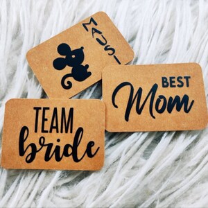 Könnte beinhalten: Drei braune Papier-Geschenk-Etiketten mit schwarzem Text. Die erste Etikette sagt "Team bride" in einer Schriftart. Die zweite Etikette sagt "Best Mom" in einer Schriftart. Die dritte Etikette hat eine schwarze Silhouette einer Maus und den Text "MADS" vertikal geschrieben.