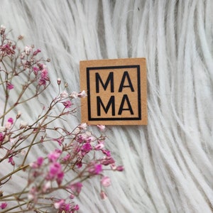 Può includere: Un blocco di legno quadrato marrone con la parola "MAMA" stampata in lettere nere.