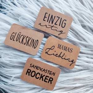 Könnte beinhalten: Vier rechteckige Kraftpapier-Etiketten mit schwarzem Text. Die Etiketten lauten: "EINZIG artig", "GLÜCKSKIND", "MAMAS Liebling" und "SANDKASTEN ROCKER". Die Etiketten sind auf einer weißen, flauschigen Oberfläche angeordnet.