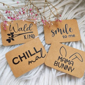 Könnte beinhalten: Vier rechteckige Kraftpapieranhänger mit schwarzem handgeschriebenem Text. Die Anhänger lesen "Wald KIND", "smile to me", "CHILL mal" und "MAMA BUNNY". Die Anhänger sind mit einem Zweig rosa Blüten dekoriert.