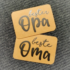 Könnte beinhalten: Zwei braune rechteckige Karten mit schwarzer Beschriftung. Die obere Karte sagt "bester Opa" und die untere Karte sagt "beste Oma".