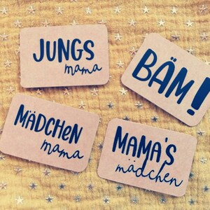 Può includere: Quattro carte marroni con testo nero su sfondo giallo con stelle dorate. Le carte dicono "Jungs mama", "Mädchen mama", "Mama's Mädchen", e "BÄM!"