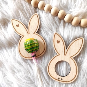May include: Two wooden bunny-shaped lollipop holders. One holder has a green and yellow lollipop in it. Both holders have a small heart cutout on the ears.