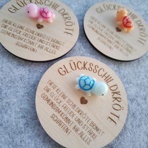 Op de afbeelding: Drie houten ronde plaquettes met de Duitse tekst "GL&Uuml;CKSSCHILDKR&Ouml;TE" en een inspirerende boodschap. Elke plaquette heeft een klein, kleurrijk schildpadfiguur. De schildpadden zijn roze, oranje en blauw.