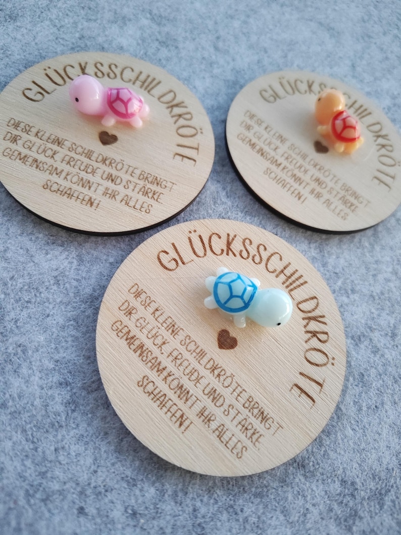 Op de afbeelding: Drie houten ronde plaquettes met de Duitse tekst "GL&Uuml;CKSSCHILDKR&Ouml;TE" en een inspirerende boodschap. Elke plaquette heeft een klein, kleurrijk schildpadfiguur. De schildpadden zijn roze, oranje en blauw.