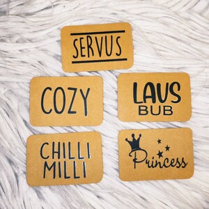 Può includere: Cinque etichette regalo in carta marrone con testo nero. Le etichette recitano "Servus", "Cozy", "Laus Bub", "Chilli Milli" e "Princess" con una corona e stelle.