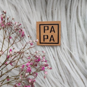 Könnte beinhalten: Ein braunes Papieretikett mit einem schwarzen quadratischen Rand und dem Text "PA PA" darin.
