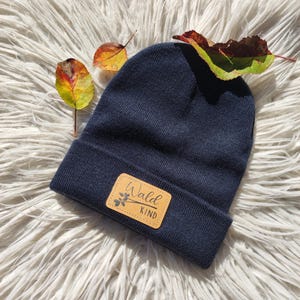 Könnte beinhalten: Eine dunkelblaue Mütze mit einem braunen Lederpatch, auf dem "Wald Kind" in schwarzen Buchstaben steht.