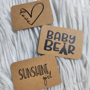Könnte beinhalten: Drei braune Geschenkanhänger aus Papier mit schwarzer Tinte. Der erste Anhänger hat ein Herz mit dem Wort "Love" darin geschrieben. Der zweite Anhänger sagt "Baby Bear" mit einer kleinen Zeltgrafik. Der dritte Anhänger sagt "Sunshine girl".