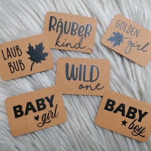 Pode incluir: Seis etiquetas de papel castanho com texto preto e imagens. As etiquetas dizem "Laub Bub" com uma folha de bordo, "Räuber kind", "Golden girl" com uma folha de bordo, "Wild one", "Baby girl" com um coração e "Baby boy" com uma estrela.