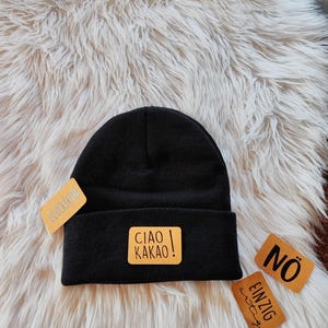 Könnte beinhalten: Schwarze Mütze mit einem gelben rechteckigen Aufnäher mit der Aufschrift "CIAO KAKAO!". Zusätzliche Etiketten mit den Wörtern "GLÜCKSKIND", "NÖ" und "EINZIG" sind ebenfalls zu sehen. Die Mütze liegt auf einer weißen, flauschigen Oberfläche.
