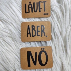 Può includere: Tre carte rettangolari marroni con testo nero. La carta superiore dice "Läuft..." La carta centrale dice "Aber." La carta inferiore dice "Nö."