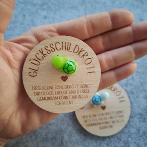 Op de afbeelding: Twee houten medaillons met de Duitse tekst "GL&Uuml;CKSSCHILDKR&Ouml;TE" en een klein schildpadfiguur. De tekst "DIESE KLEINE SCHILDKR&Ouml;TE BRINGT DIR GL&Uuml;CK, FREUDE UND ST&Auml;RKE GEMEINSAM K&Ouml;NNT IHR ALLES SCHAFFEN!" is ook zichtbaar.