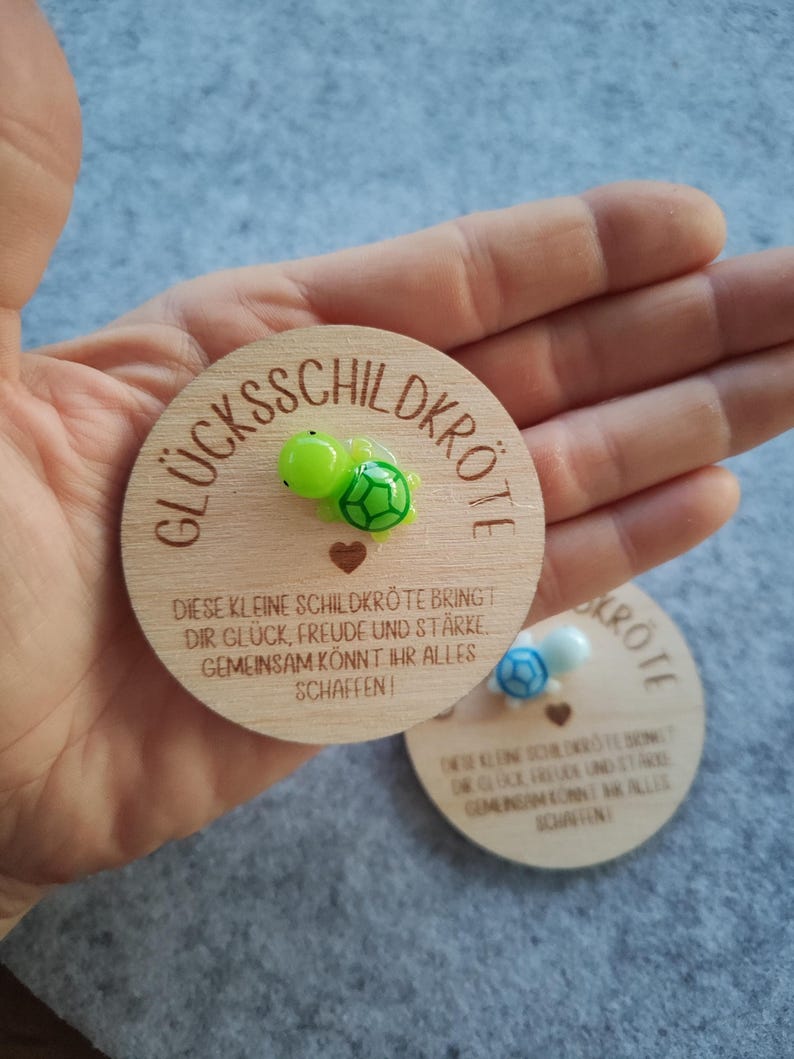 Op de afbeelding: Twee houten medaillons met de Duitse tekst "GL&Uuml;CKSSCHILDKR&Ouml;TE" en een klein schildpadfiguur. De tekst "DIESE KLEINE SCHILDKR&Ouml;TE BRINGT DIR GL&Uuml;CK, FREUDE UND ST&Auml;RKE GEMEINSAM K&Ouml;NNT IHR ALLES SCHAFFEN!" is ook zichtbaar.
