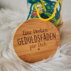 Könnte beinhalten: Ein runder Holzdeckel mit dem eingravierten Text "Eine Portion Geduldfäden für Dich". Der Deckel liegt auf einer weißen, flauschigen Oberfläche.