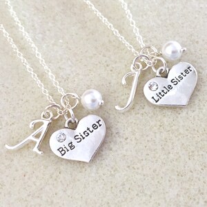 May include: Two silver necklaces with heart pendants. One pendant says "Big Sister" and the other says "Little Sister". Each necklace has a pearl charm and a letter charm.