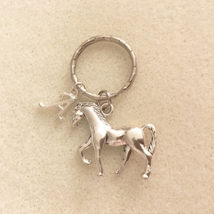 May include: Silver horse keychain with a letter A charm. The horse is standing on all four legs with its head turned to the side. The keychain is attached to a silver key ring.