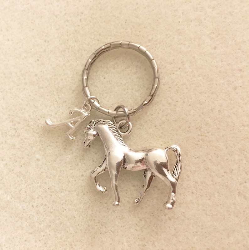 May include: Silver horse keychain with a letter A charm. The horse is standing on all four legs with its head turned to the side. The keychain is attached to a silver key ring.