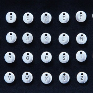 May include: A set of 26 silver-toned metal charms, each featuring a different letter of the alphabet. The charms are circular and have a small loop at the top for attaching to a necklace, bracelet, or other jewelry.