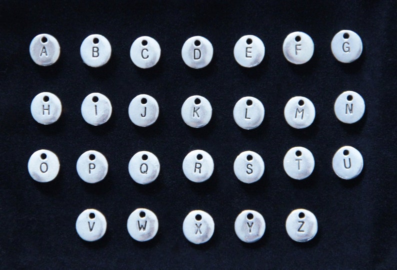 May include: A set of 26 silver-toned metal charms, each featuring a different letter of the alphabet. The charms are circular and have a small loop at the top for attaching to a necklace, bracelet, or other jewelry.