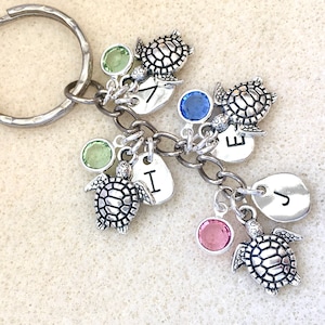 May include: A silver keychain with a silver key ring and multiple charms. The charms include four silver turtle charms, four silver heart charms with the letters J, I, E, and V, and four crystal charms in green, blue, pink, and green.