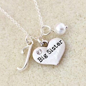 May include: Silver necklace with a heart charm that says "Big Sister" and a letter "J" charm. The necklace also has a small pearl charm.