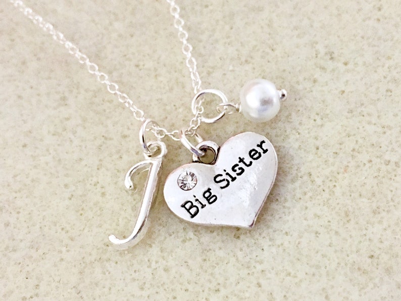 May include: Silver necklace with a heart charm that says "Big Sister" and a letter "J" charm. The necklace also has a small pearl charm.