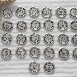 Może przedstawiać: Zestaw 26 srebrzystych metalowych zawieszek, każda z inną literą alfabetu. Zawieszki są okrągłe i mają wypukły wz&oacute;r liter.