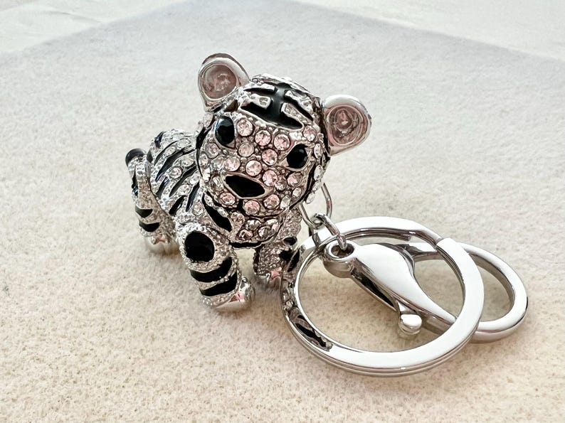 May include: A silver and black jeweled tiger keychain with a silver key ring. The tiger is covered in clear and pink rhinestones.