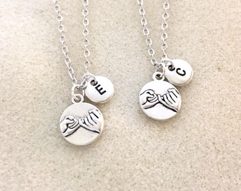 May include: Two silver necklaces with a charm of two hands making a pinky promise and a small round charm with a letter on it. The necklaces are on a white background.