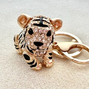 May include: Gold-tone tiger keychain with black and white stripes and clear rhinestones. The tiger is sitting on a gold-tone key ring.