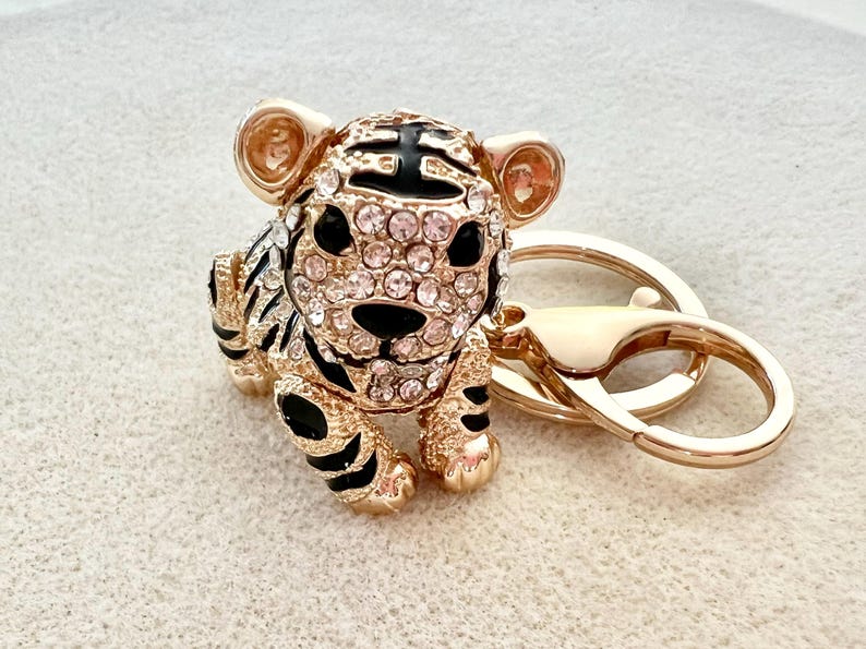 May include: Gold-tone tiger keychain with black and white stripes and clear rhinestones. The tiger is sitting on a gold-tone key ring.