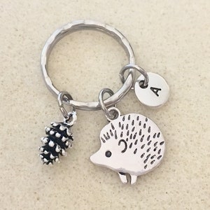 May include: Silver keychain with a hedgehog charm, a pine cone charm, and a silver disc charm with the letter 'A' on it.