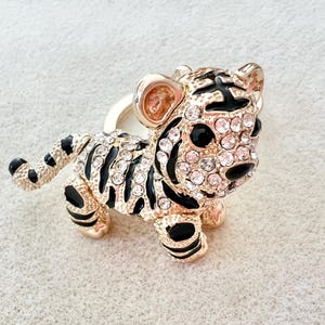 May include: A gold-tone tiger figurine with black stripes and clear rhinestones. The figurine is standing on a white surface.