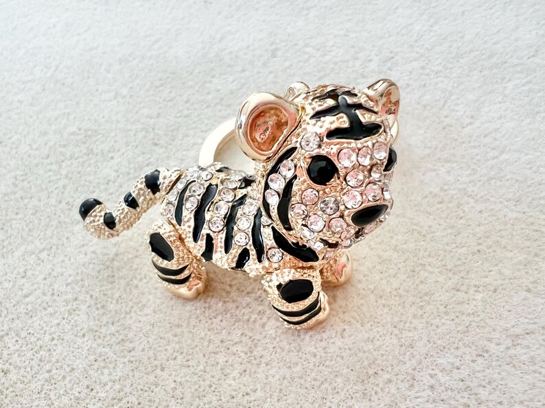 May include: A gold-tone tiger figurine with black stripes and clear rhinestones. The figurine is standing on a white surface.