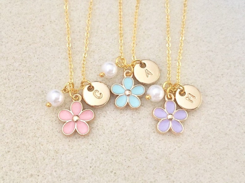 Pu&ograve; includere: Tre collane d'oro con catene delicate, ciascuna con una piccola perla, un ciondolo rotondo con una lettera e un ciondolo a forma di fiore color pastello. I ciondoli sono rosa, blu e viola.
