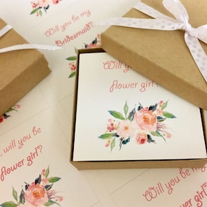 May include: Kraft paper boxes with white ribbon and floral cards. The cards read "Will you be my Bridesmaid?" and "Will you be my flower girl?" in pink script, with watercolor floral designs.