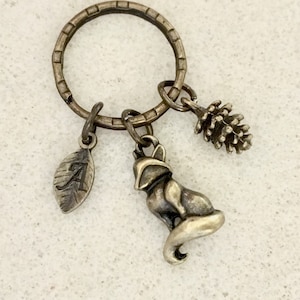 May include: Silver-toned keychain with a ring holding three charms: a leaf with the letter "A", a fox, and a pinecone. The charms are detailed and have an antique finish. The keychain is likely made of metal.