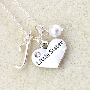 May include: Silver necklace with a heart charm that says "Little Sister" and a letter J charm. The necklace also has a small pearl charm.