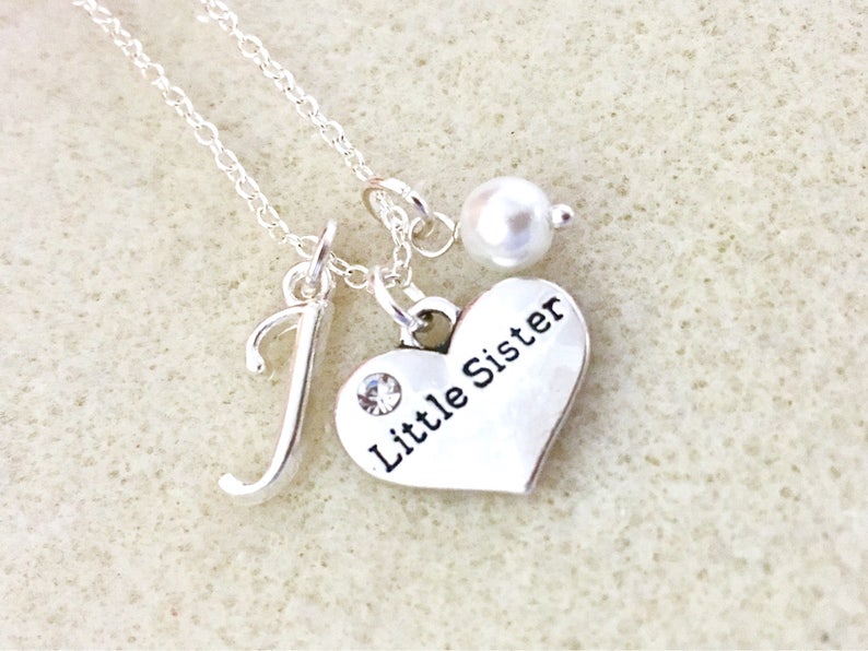 May include: Silver necklace with a heart charm that says "Little Sister" and a letter J charm. The necklace also has a small pearl charm.