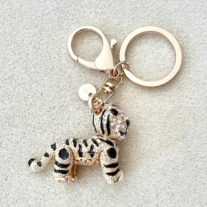 May include: Gold-tone key chain with a small, jeweled tiger charm. The tiger is black and white with clear rhinestones.