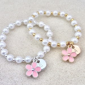 May include: Two pearl bracelets with flower charms. One bracelet has gold beads and the other has silver beads. The charms are pink with a silver and gold finish. The charms have the letters "A" and "LL" on them.