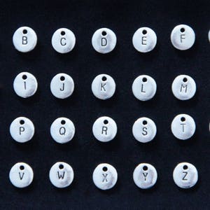 May include: A set of 26 silver metal alphabet letter charms. Each charm is round and has a single letter engraved on it. The letters are in uppercase and are arranged in alphabetical order.